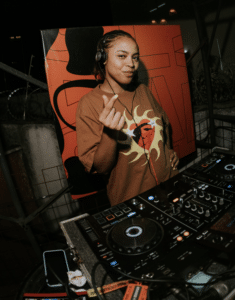 A DJ wearing headphones stands behind a mixing console, holding one hand up and making a finger heart gesture, surrounded by vibrant artwork—perfect for music lovers and unique gifts.
